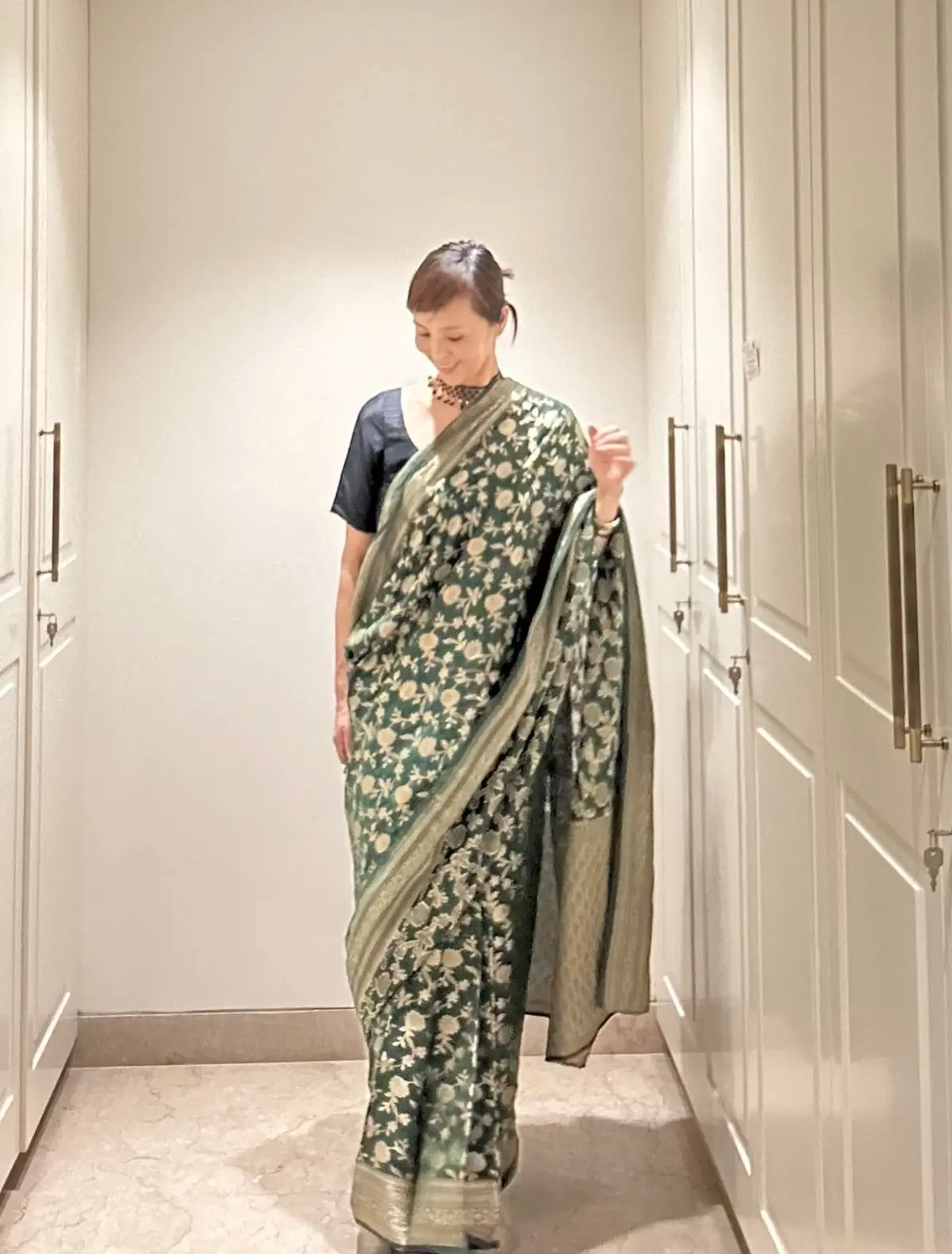 緑色の花柄のサリーを着た女性が、白い収納棚が並ぶ廊下を歩いている。天井には扇風機があり、廊下の奥にスポットライトが見える。