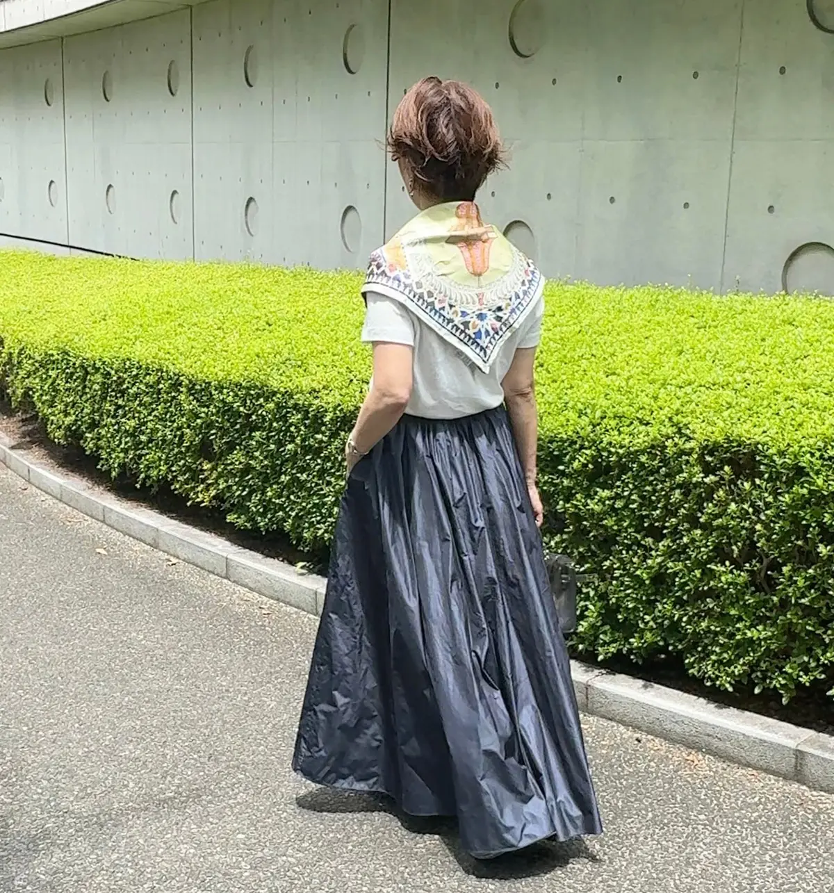 女性が後ろ姿で立っている写真。白いTシャツに、カラフルなスカーフを巻いている。紺色のフレアスカートを着用し、緑の植栽の前でポーズをとっている。背景には、穴の開いたコンクリートの壁がある。