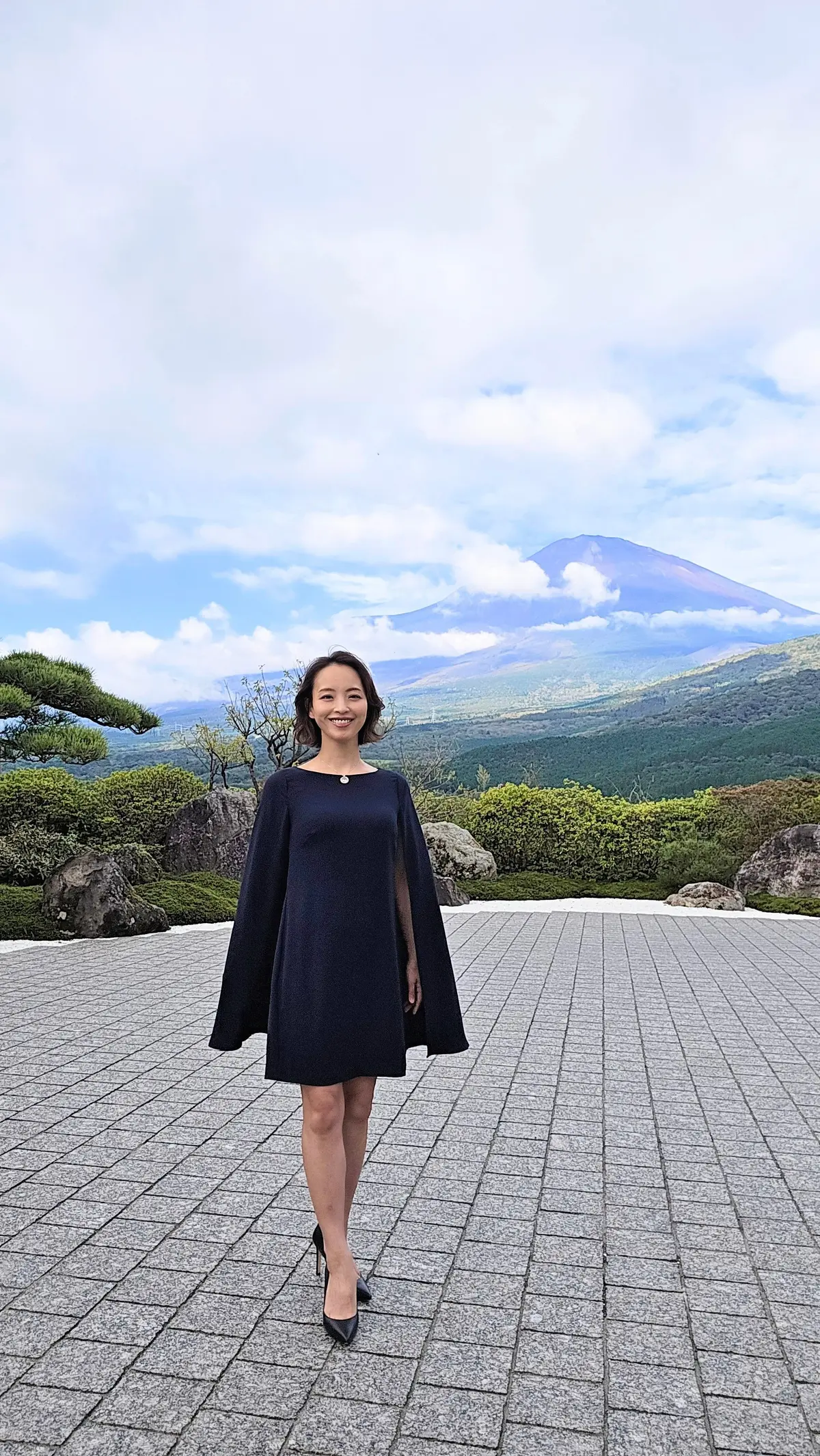 黒いケープ付きのワンピースを着た女性が、背景に富士山を背に立っている。女性は笑顔で、黒いパンプスを履いている。石畳の広場にいる。