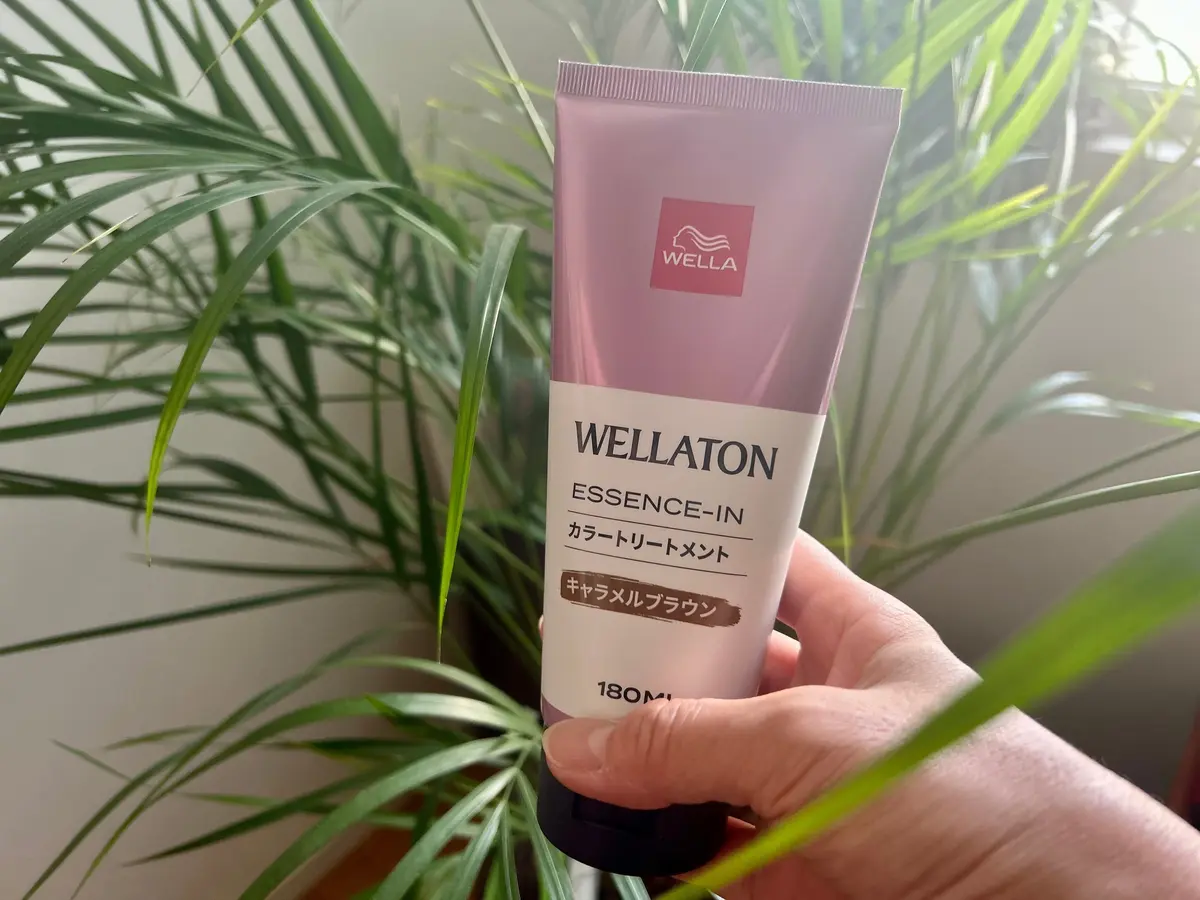 Wellaのピンク色のヘアケア製品「Wellaton エッセンス・イン クラートリートメントメント」を手に持つ手。背景には緑の植物。