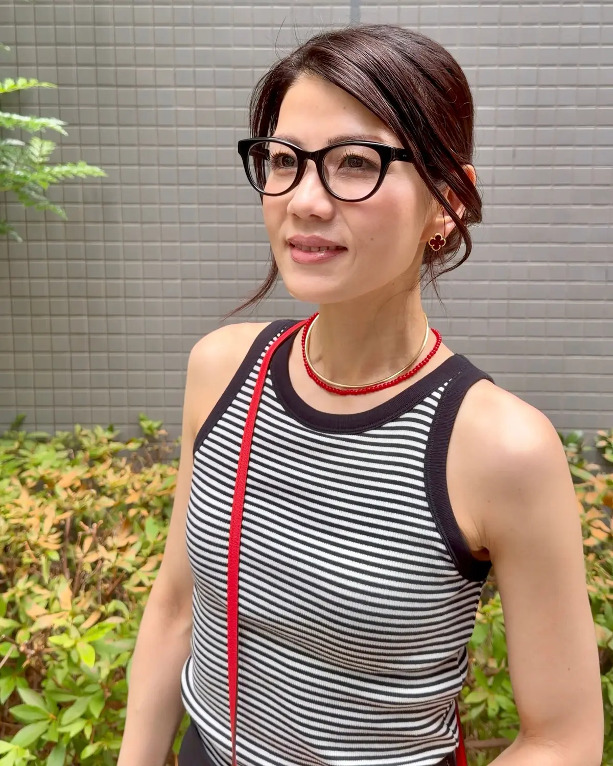 黒縁メガネをかけた女性が、横を向いて微笑んでいる。黒と白のボーダーのノースリーブに、赤いネックレスとイヤリングを着用。背景にはグレーの壁と緑の植物がある。