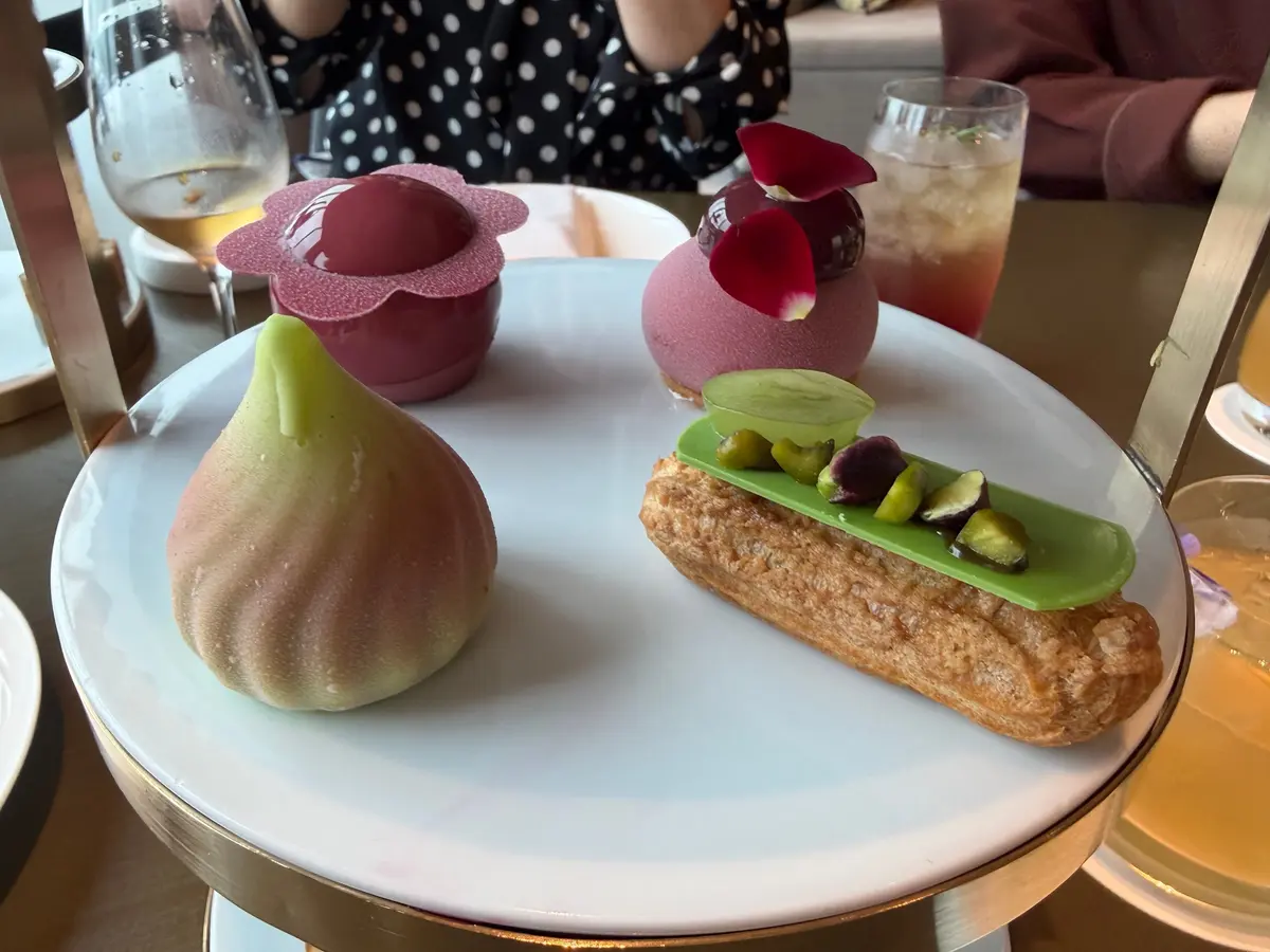 白い円盤に、赤紫色の花形スイーツ、丸いムース、梨の形をした焼き菓子、緑色のエクレアが並んでいる。背景には飲み物と人物がぼやけて写っている。