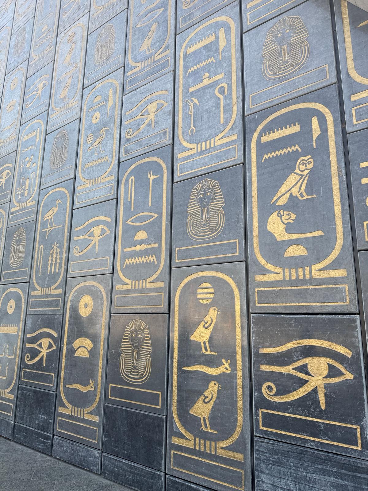黒色のタイル壁に、金色の古代エジプトのヒエログリフが規則的に配置されている。縦長の長方形の象形文字が並び、鳥や人、目などのモチーフが見られる。