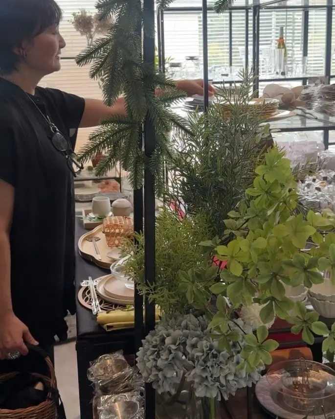 黒いシャツを着た女性が、クリスマスデコレーションされたテーブルの植物に手を伸ばしている。テーブルには皿やカトラリー、キャンドルなどが並び、背景には窓が見える。