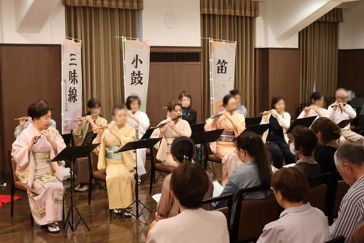 和服姿の女性たちが座って篠笛を演奏し、観客が前列に座っている室内コンサート。