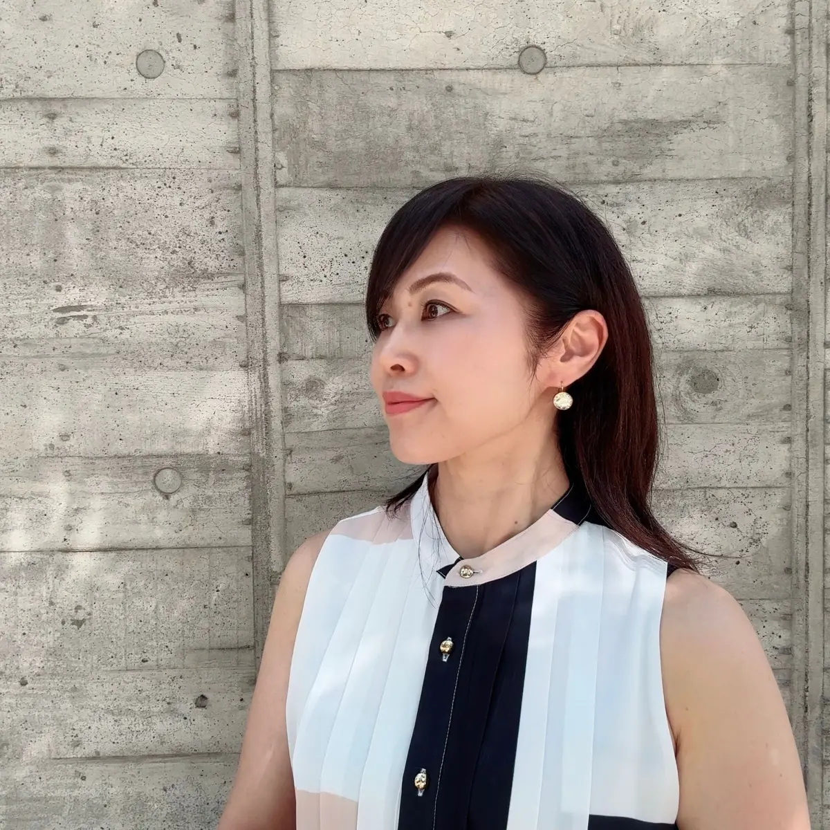 黒髪の女性が、白い装飾が施された黒と白のトップスを着て、横を向いて微笑んでいる。背景はグレーのコンクリート壁。耳には控えめなイヤリングを着用。