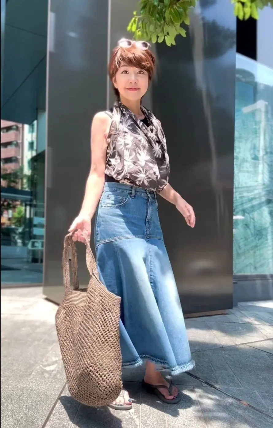 女性がデニムスカートと花柄トップスを着て、屋外でカゴバッグを持っています。頭頂にサングラスをかけ、モダンなガラスの建物前に立っています。