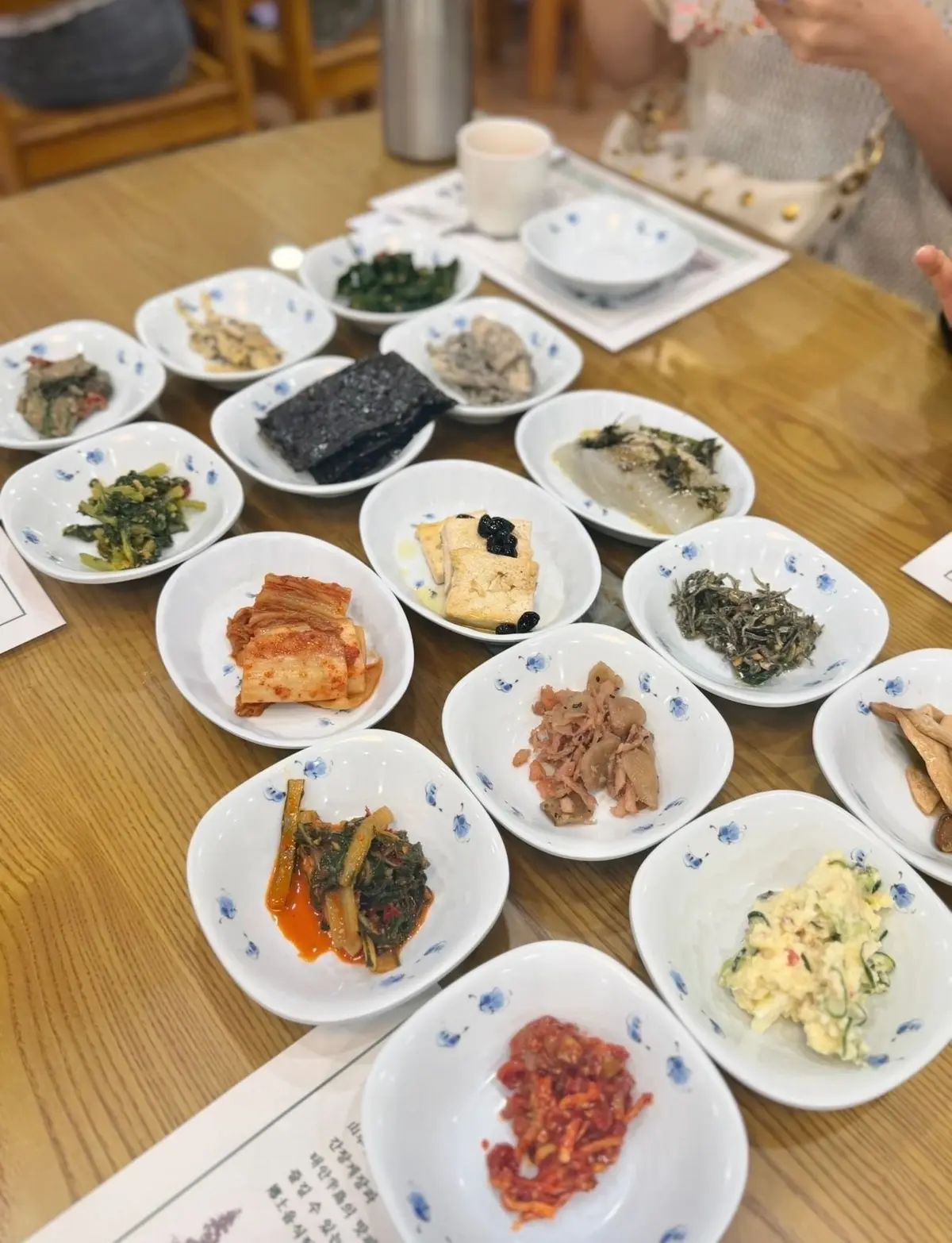木製のテーブルに並べられた、白地の小皿に盛り付けられた韓国のキムチの盛り合わせ。様々な色と種類のキムチが、整然と並んでいる。