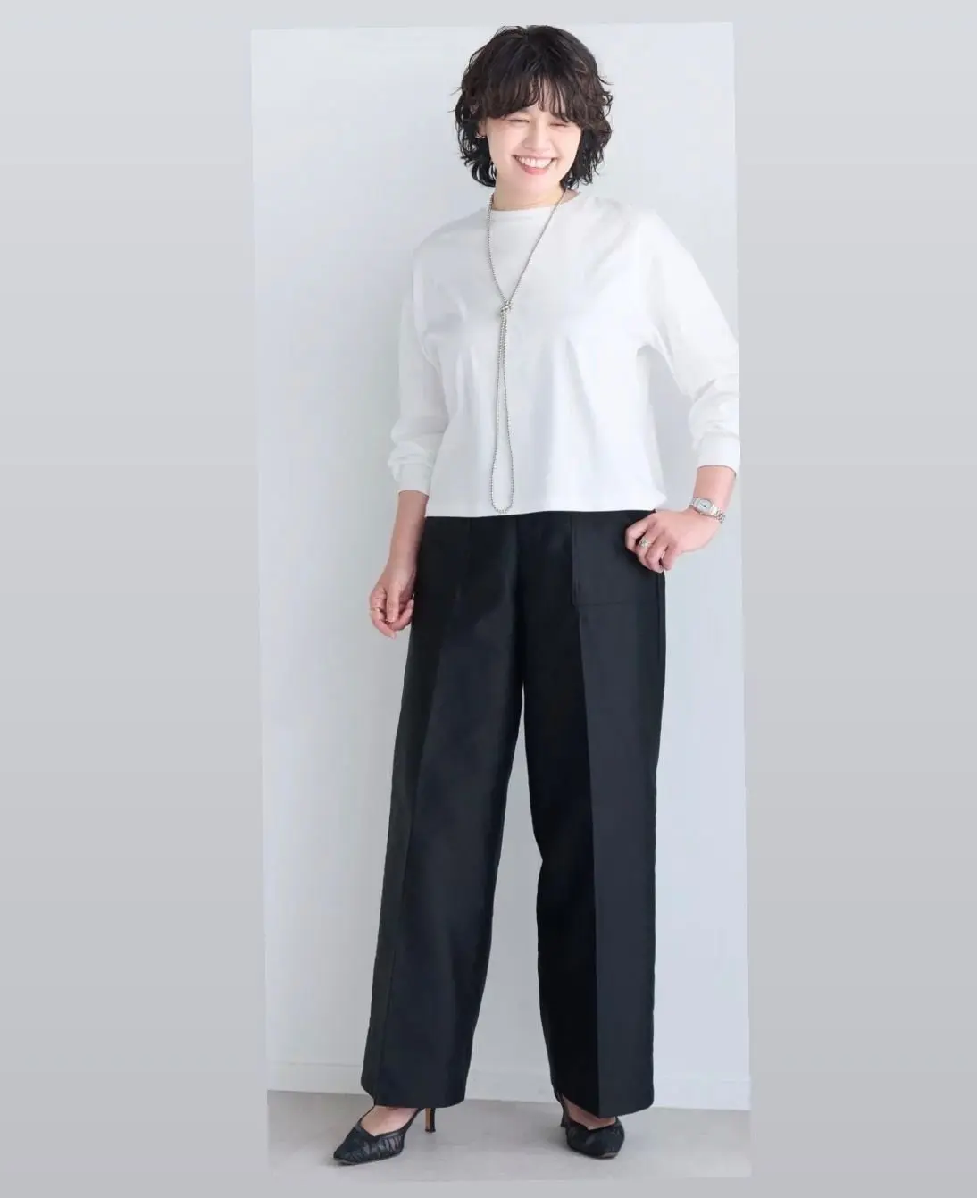 白い長袖カットソーに黒いワイドパンツを合わせた女性。茶色の短髪で、笑顔を見せている。シルバーのネックレスと腕時計を着用。背景は淡いグレー。
