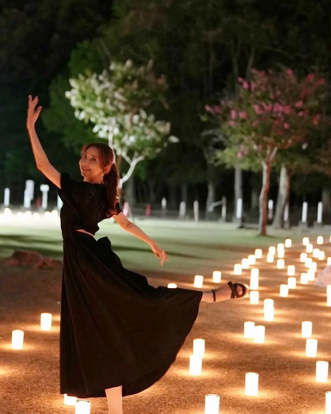 夜の公園で、黒いワンピースを着た女性が右手を上げて踊っている。周りには白いキャンドルが点灯し、ロマンチックな雰囲気を醸し出している。