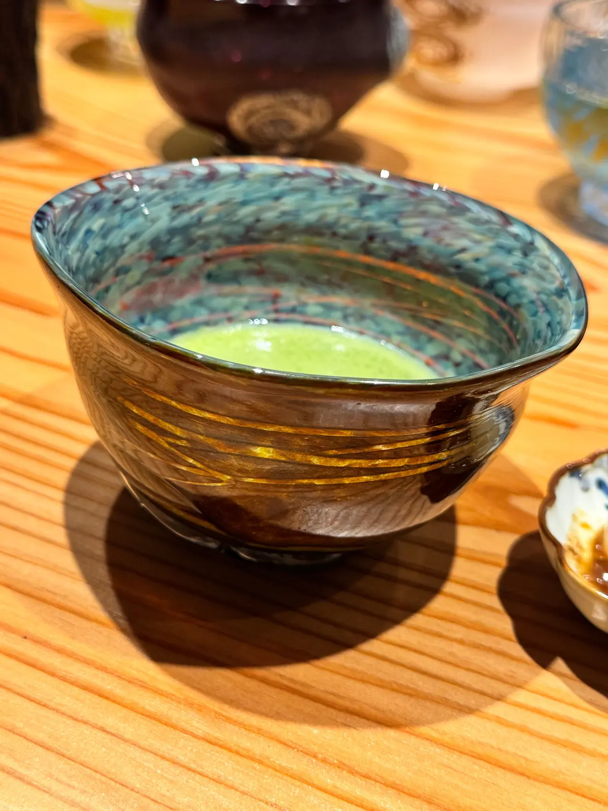 茶碗に緑色の抹茶が注がれている。茶碗は青と茶色の模様が入り、木製のテーブルに置かれている。背景には他の茶碗が見える。