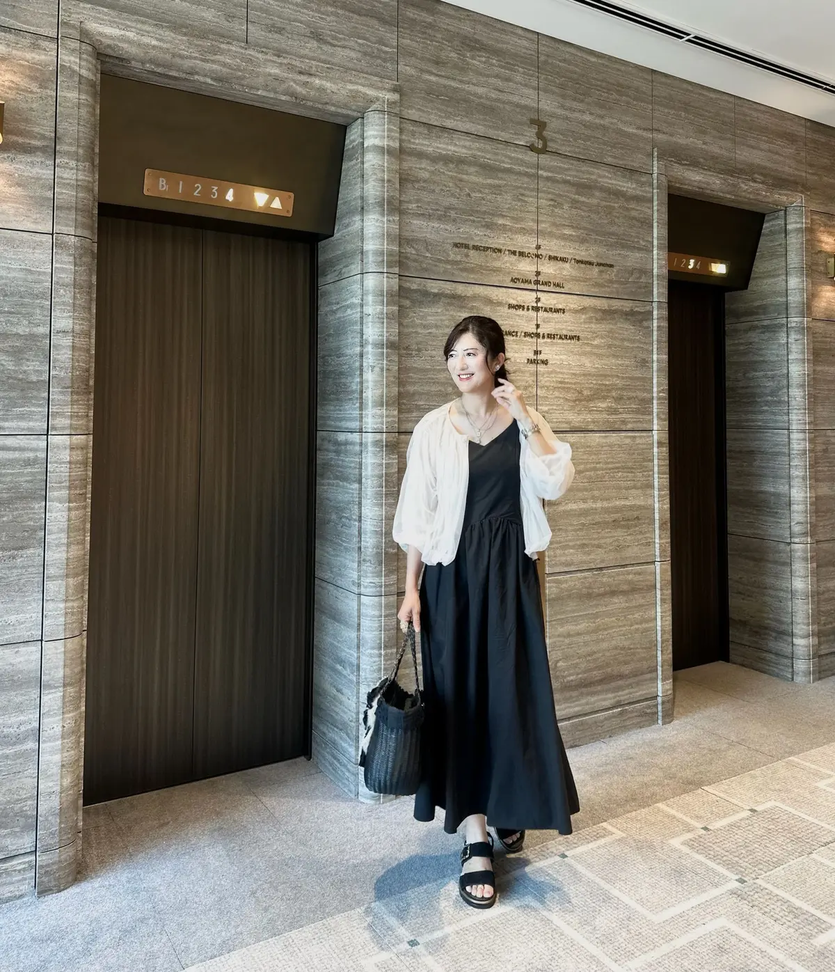黒いキャミソールワンピースに白のトップスを羽織った女性が、エレベーター前で微笑んでいる全身写真。黒いバッグを持ち、黒いサンダルを履いている。背景はグレーの石材で覆われた壁。