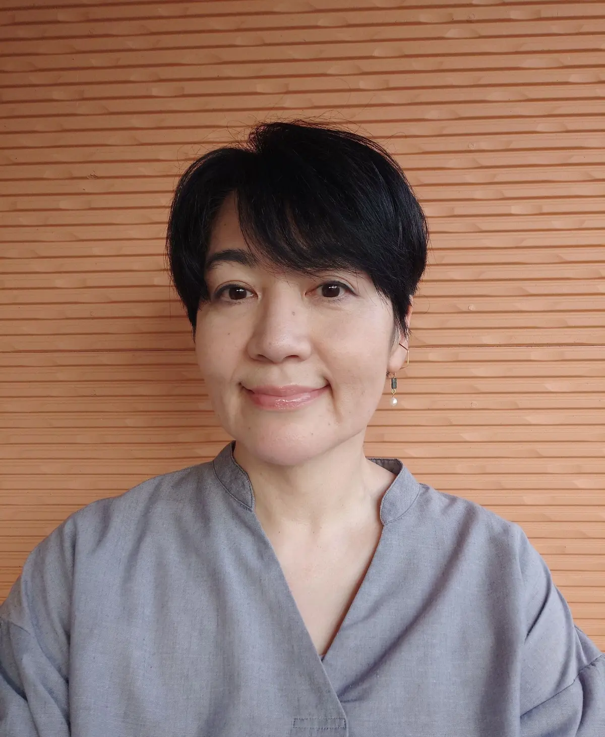 女性が淡い灰色の服を着て、木製の壁前に立っている。短い黒髪で、耳飾りを身につけ、ほほ笑んでいる。