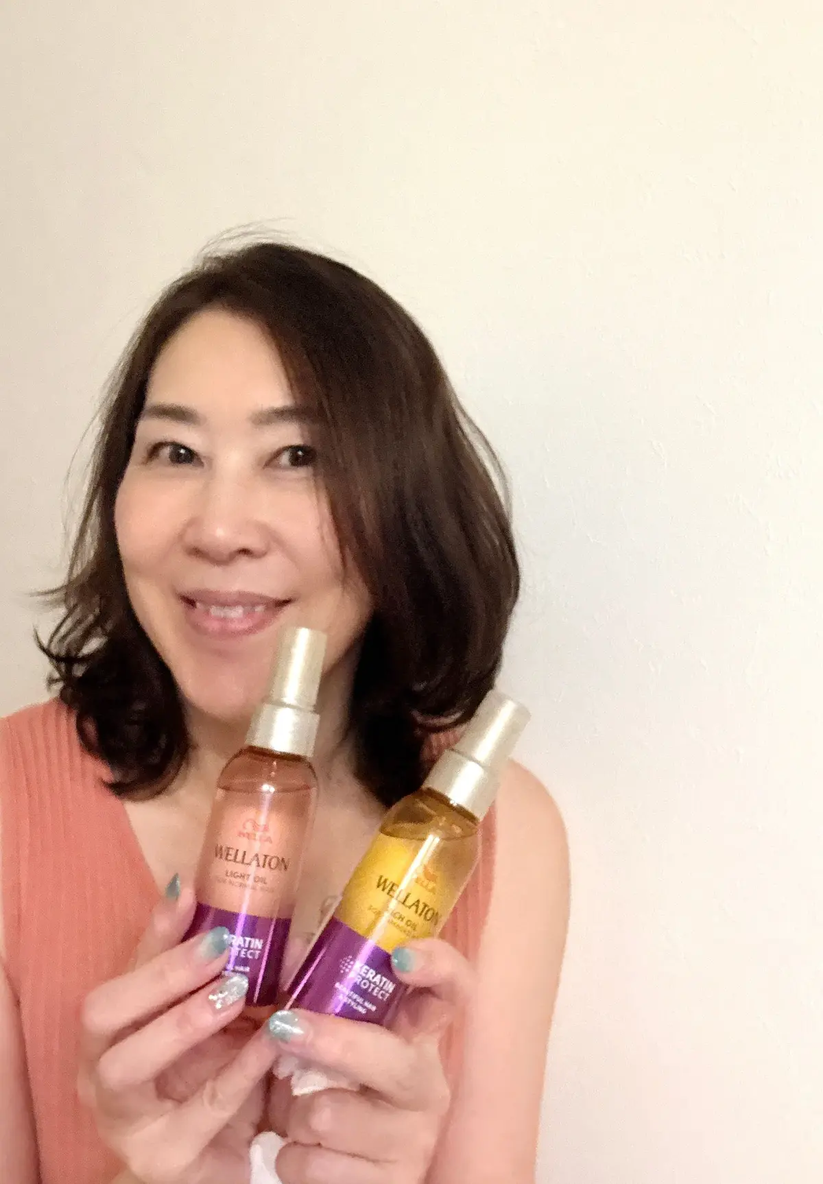 笑顔の女性が、ウェラTON（WellaTON）のヘアオイル2本を持ち、カメラ目線でこちらを見ている。茶色のボブヘアで、オレンジ色のトップスを着用。背景は壁。
