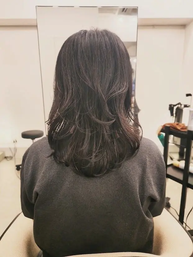 肩下丈のダークブラウンの長い髪を後ろから見た写真。ゆるやかなウェーブがかかり、光沢がある。背景には鏡と美容室の備品が見える。