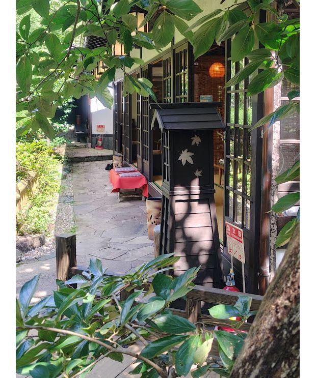木々の葉が茂る中、石畳の通路が続く。右手に格子窓の建物があり、赤いテーブルクロスが置かれたテーブルと、犬の置物が置かれている。