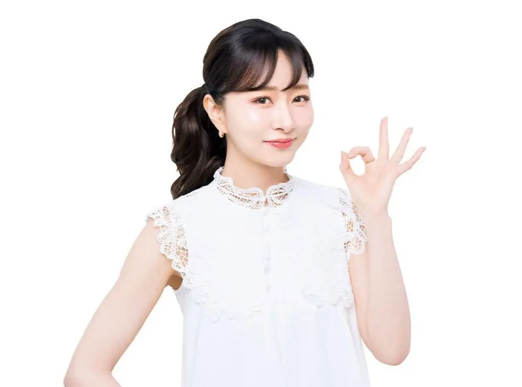 白いブラウスを着た若いアジア系の女性が、白い背景で右手を挙げて「OK」サインをしている。茶色のボブヘアを後ろで束ね、微笑んでいる。