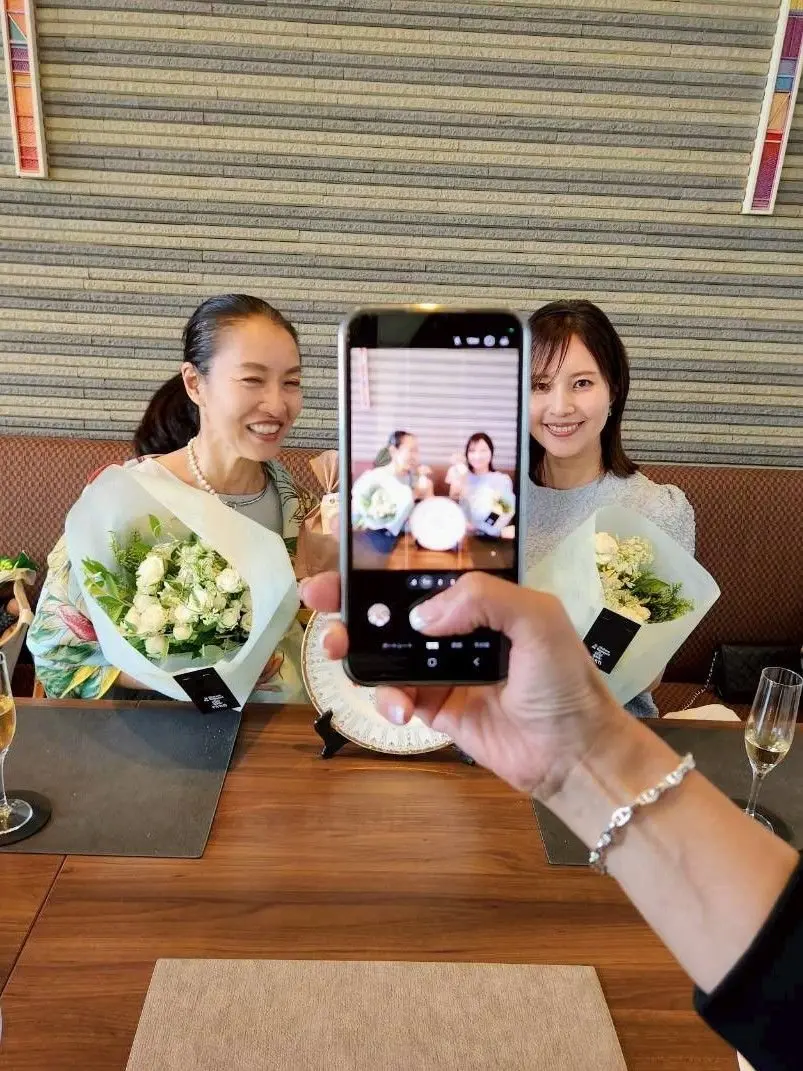 二人の女性が白い花のブーケを持ち、テーブルに座っている。手前で誰かがスマホで写真を撮っている。