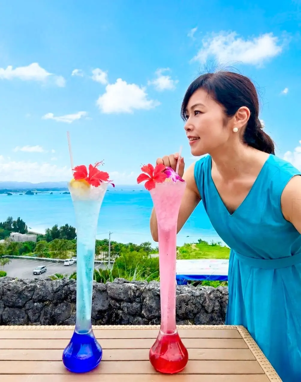 青いドレスの女性が、海の景色を背景に2つのカラフルなカクテルを手に持つ。左のカクテルは青と白の層で赤い花を飾り、右はピンクの層で赤と白の花を飾る。
