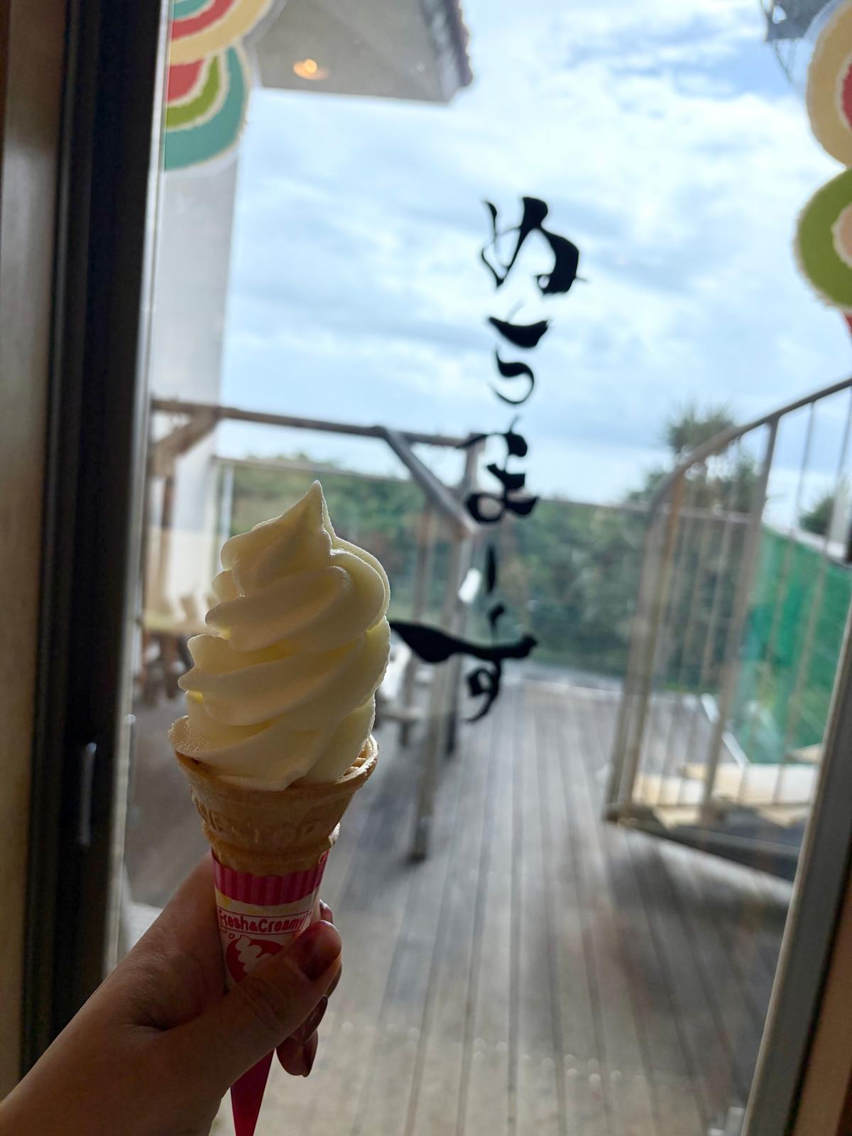 ソフトクリームを手に持ち、背景に木製の橋と緑の風景が広がる。ソフトクリームは黄色で、ワッフルコーンに入っている。手に持っている容器には「itta-Creamy」のロゴマークがある。