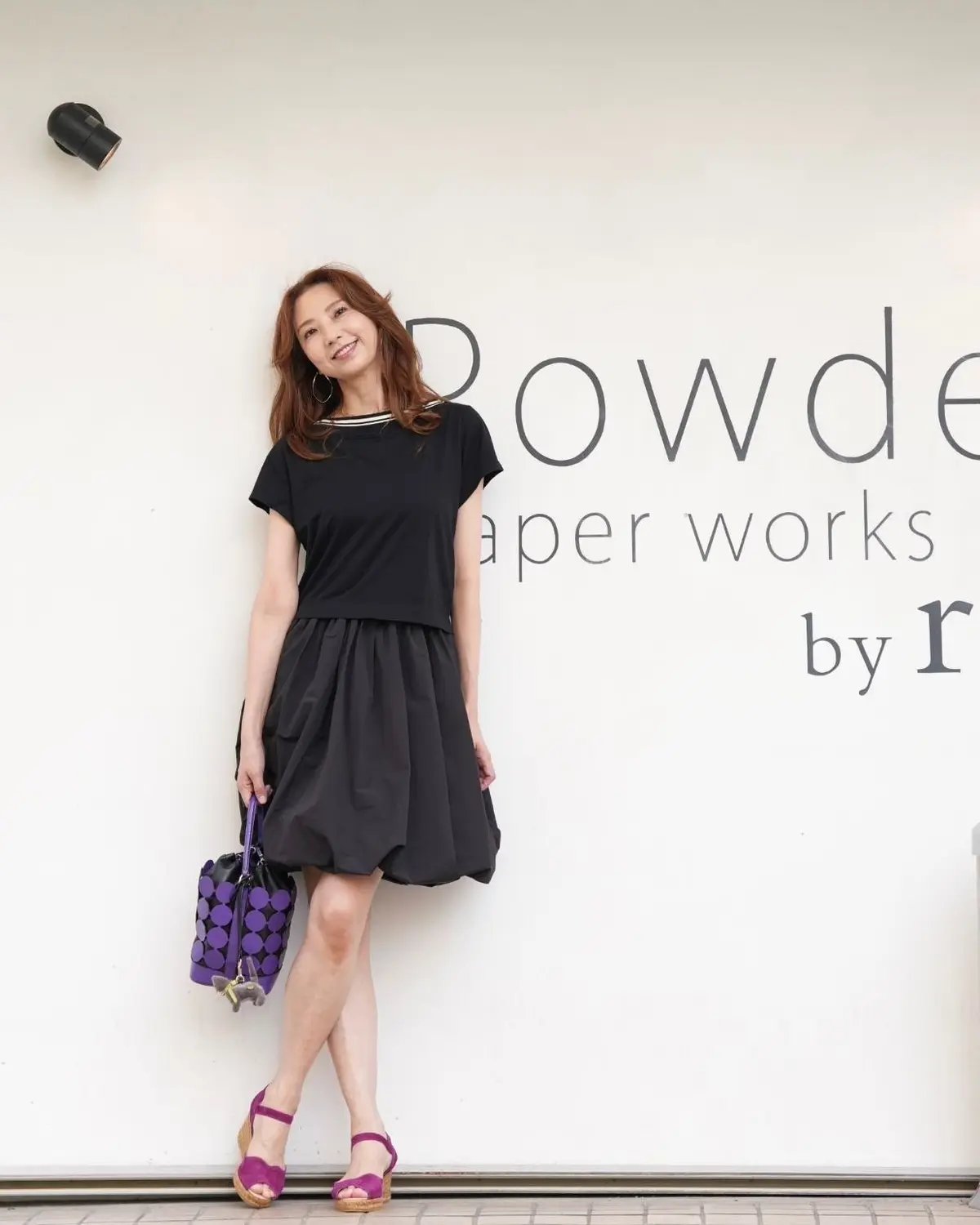 黒いトップスとスカートを着用した女性が、Rowdeの店舗の前でポーズをとっている。紫色のハンドバッグとサンダルを身につけている。