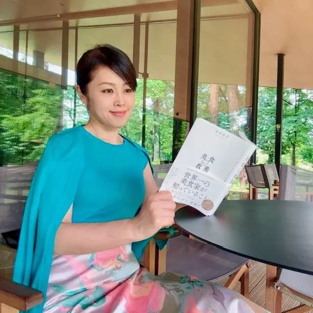 女性が青いカーディガンで窓辺のテーブルに座り、本を手に持つ。背景には緑の木々が見える。