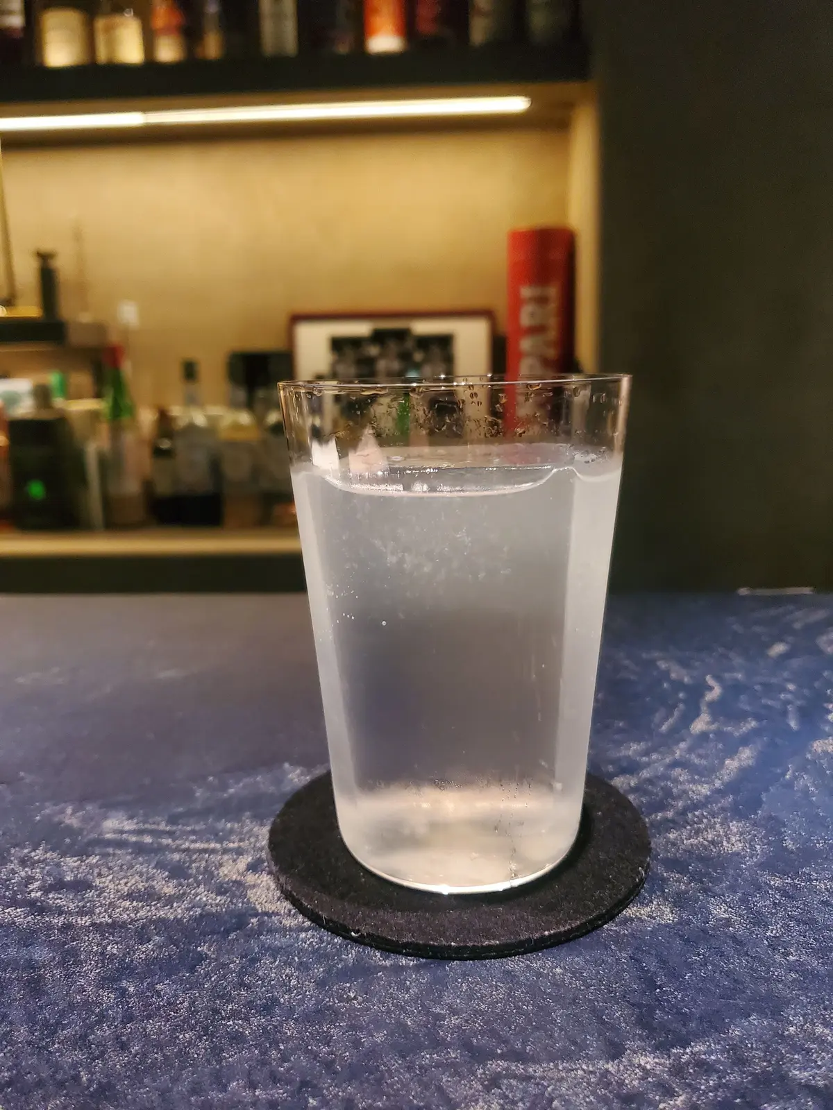 ガラスカップに水が注がれ、黒いコースターの上に置かれている。背景にはバーカウンターとさまざまなボトルが並んでいる。