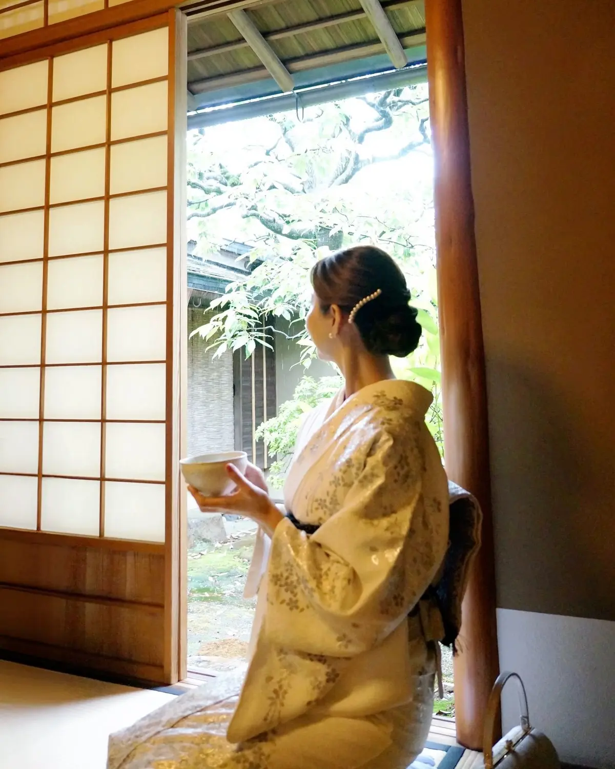 和服を着た女性が、木製の襖の開いた部屋から屋外の庭園を眺め、白い茶碗を持っている