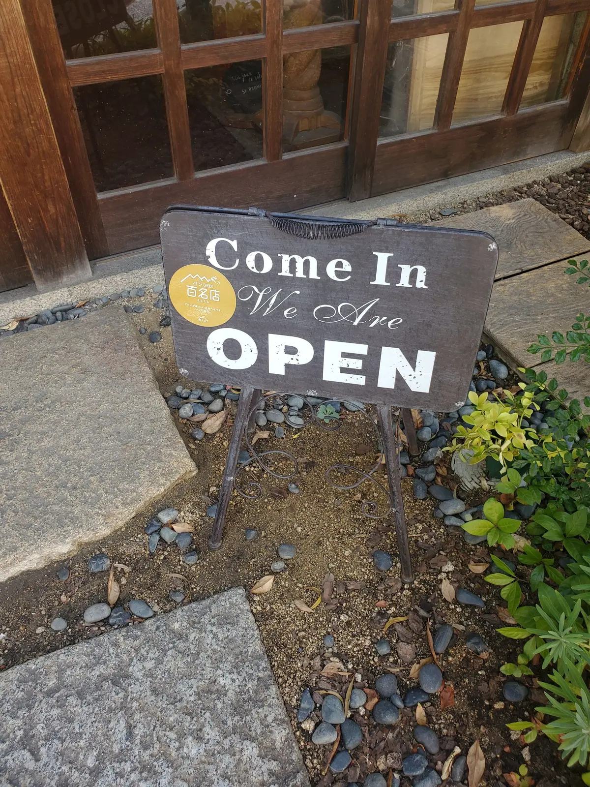 木製のイーゼルに立てられた「Come In We Are OPEN」と書かれたグレーの看板。看板には黄色いステッカーが貼られており、背景には石畳の道と植栽が見える。