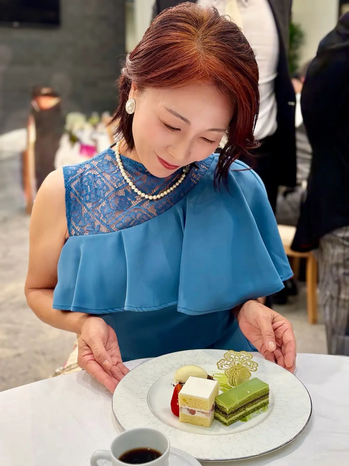 青いレースのドレスを着た女性が、花の装飾が施された白いプレート上のデザートを眺めている。プレートには緑のモンブラン、クリーム入りケーキ、フルーツが並び、コーヒーカップが置かれている。