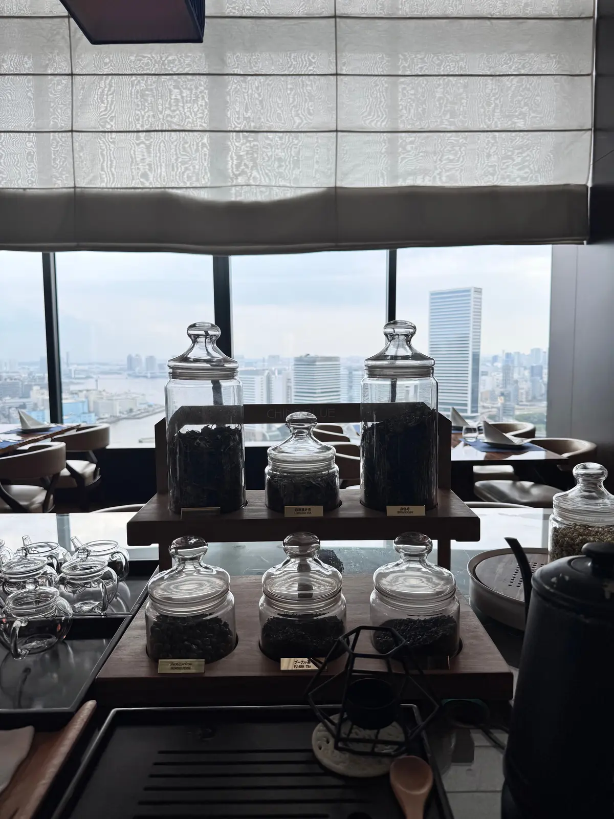 窓から都市の景色が見える、茶葉の品揃えが並んだティーバー。木製の台にガラス瓶に入った茶葉が置かれ、背景には高層ビル群が広がる。