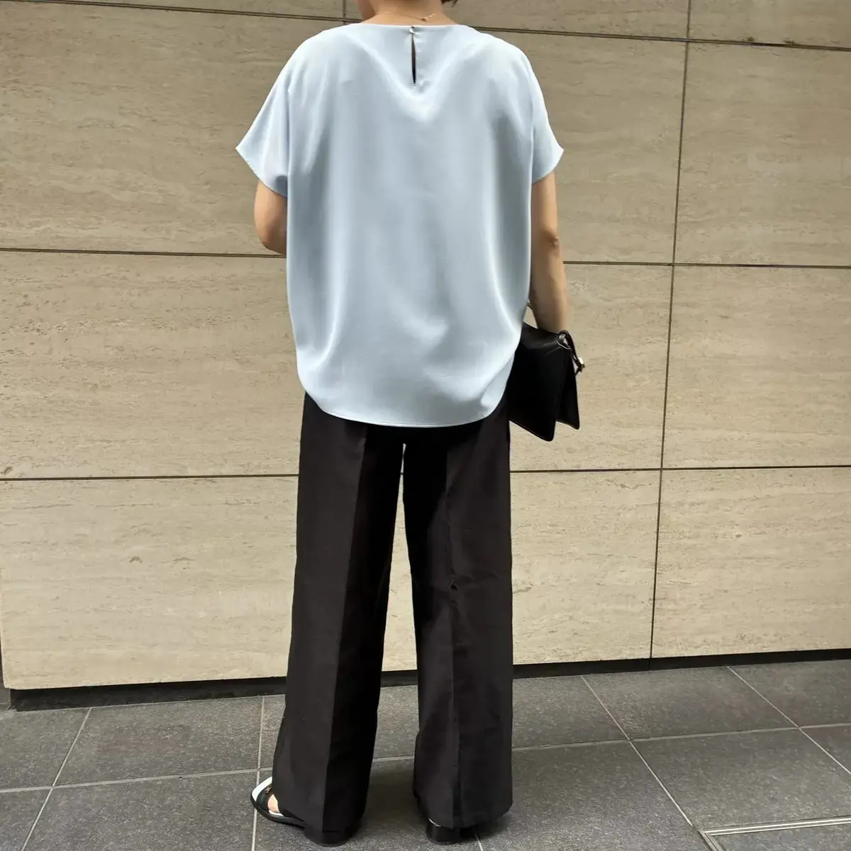 淡い水色のシルク素材の半袖トップスと黒のワイドパンツを着用した人。後ろ姿で、黒いバッグを肩に掛けている。背景はベージュ色のタイル壁。