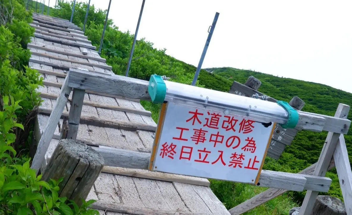 木製の階段に「工事中の為停止」と赤文字で書かれた看板と、黄色の注意看板が設置された山間部