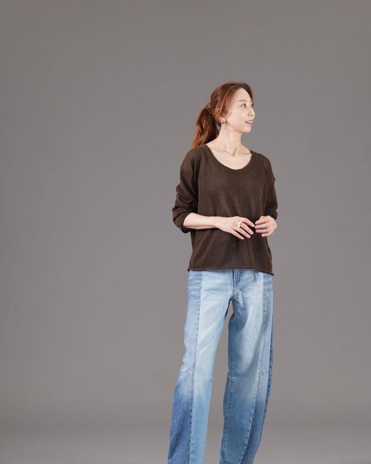 ブラウンのニットトップスと、左右に異なる色のデニムを合わせたパンツを着用した女性が、右斜め前に向かって立っている全身写真。赤茶色の髪を後ろでまとめている。背景はグレー。