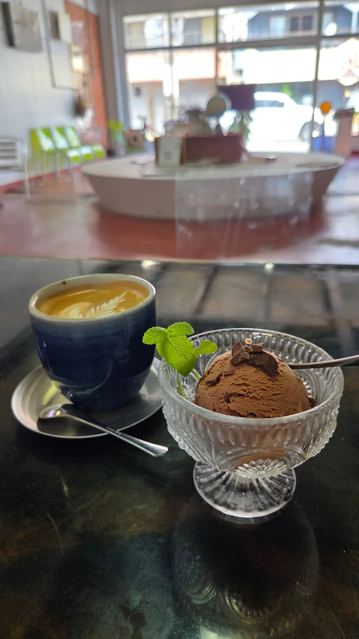 青いカップに入ったカフェラテと、ガラスの器に入ったチョコレートアイスクリーム。アイスクリームにはミントの葉とチョコレートのトッピングが添えられている。背景には窓から光が差し込み、カフェの様子が見える。