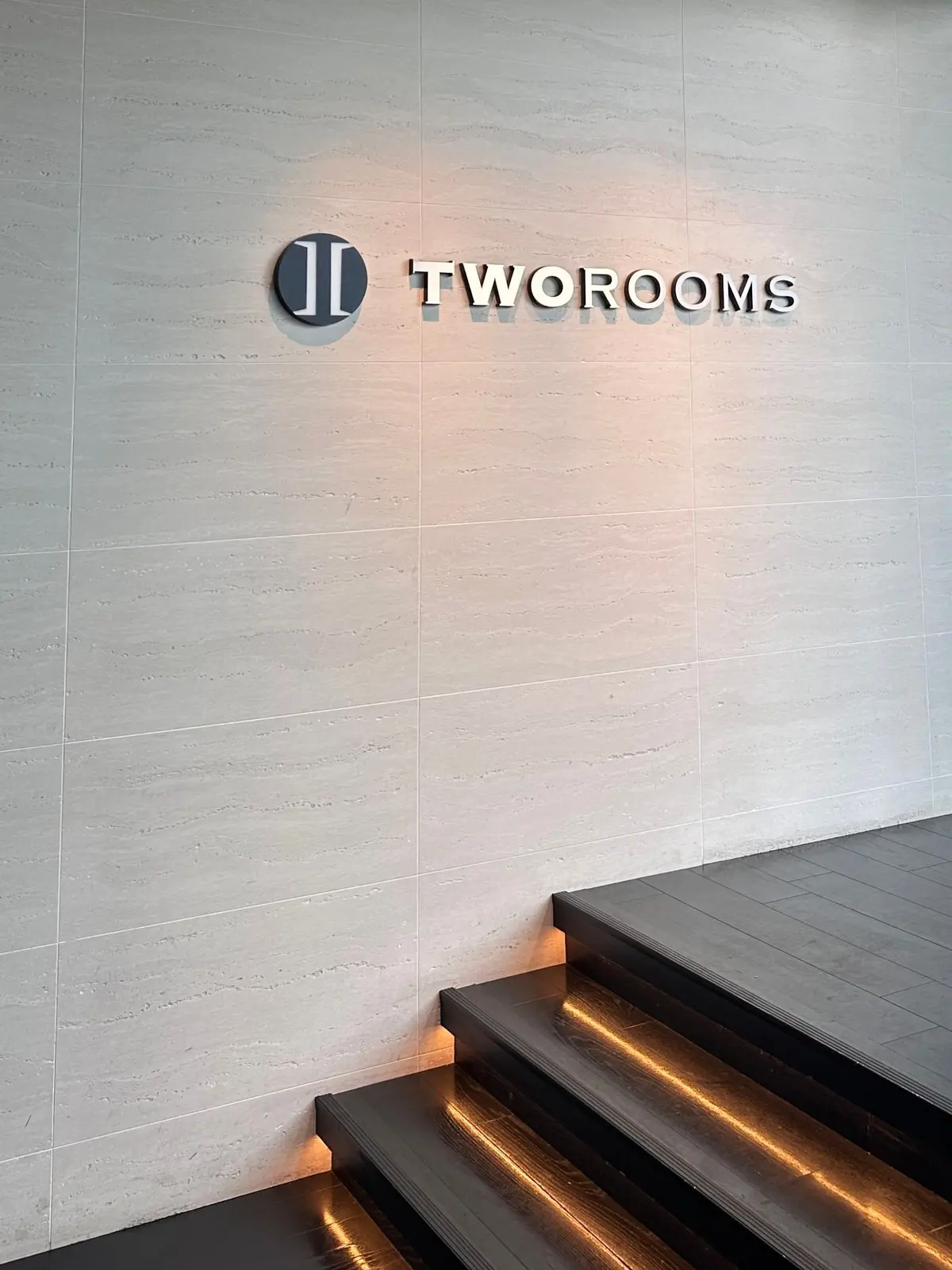 白地のタイル壁に、グレーの立体文字で「TWO ROOMS」のロゴと店名が配置されている。その下に、幅の狭い階段が設置され、照明が内照されている。