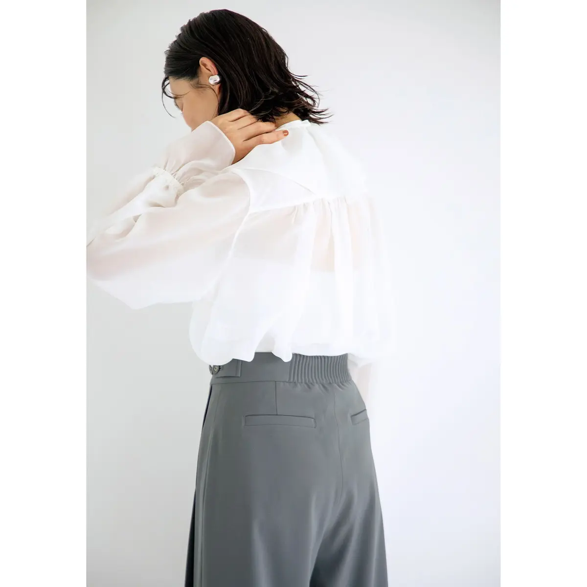 白いフリルシャツにグレーのワイドパンツを合わせた女性。笑顔でこちらを見ている。シルバーのハンドバッグを持つ。背景は白。