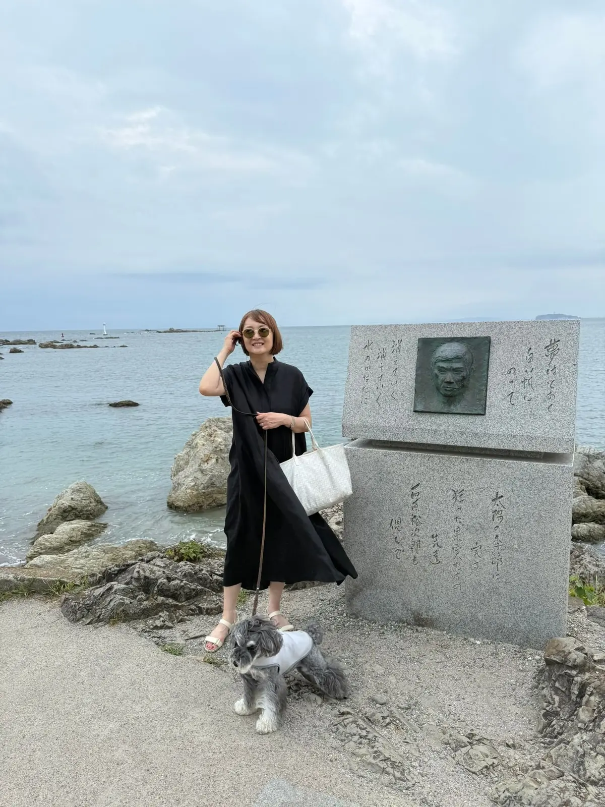 海岸の記念碑を背景に立つ女性と犬。女性は黒いワンピースにサングラスをかけ、右手にバッグを持っている。小犬は記念碑の足元に座り、こちらを見ている。背景には青い海と空が広がり、遠くに島が見える。