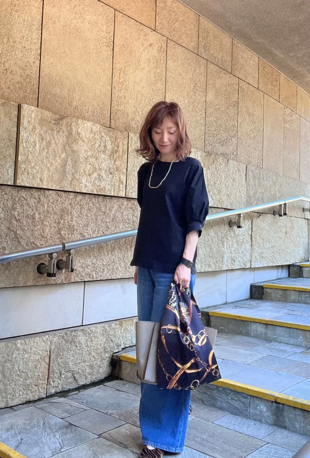 黒いトップスにデニムを合わせた女性が、階段でこちらを見る。茶色のショートヘアで、首にはゴールドのネックレスを着用。手にHermèsの柄入りのバッグを持っている。背景はベージュの石積みの壁。