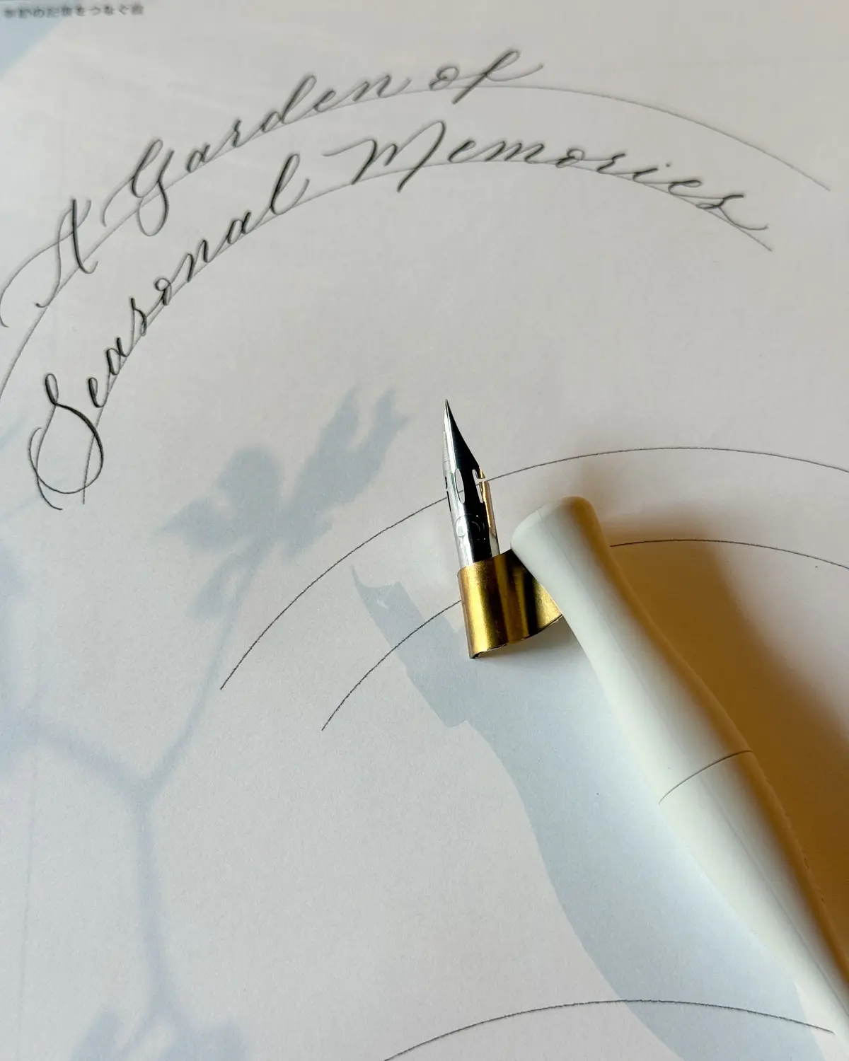 白い万年筆が紙に書かれた「A Garden of Seasonal Memories」の文字に触れている様子。背景には植物の影が映り、円弧のガイド線が引かれている。