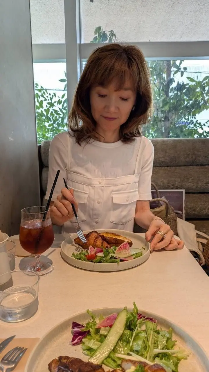 レストランで食事をする女性。白いシャツを着て、サラダと飲み物を楽しんでいる。窓から緑の植物が見える。