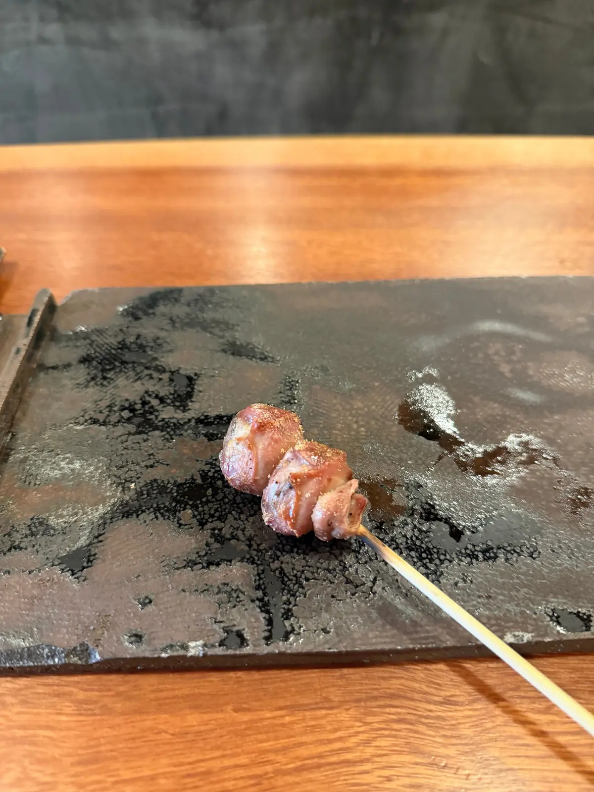 串に刺さった豚肉が、黒い石板の上にある。豚肉は表面が焼けており、やや丸みを帯びた形をしている。