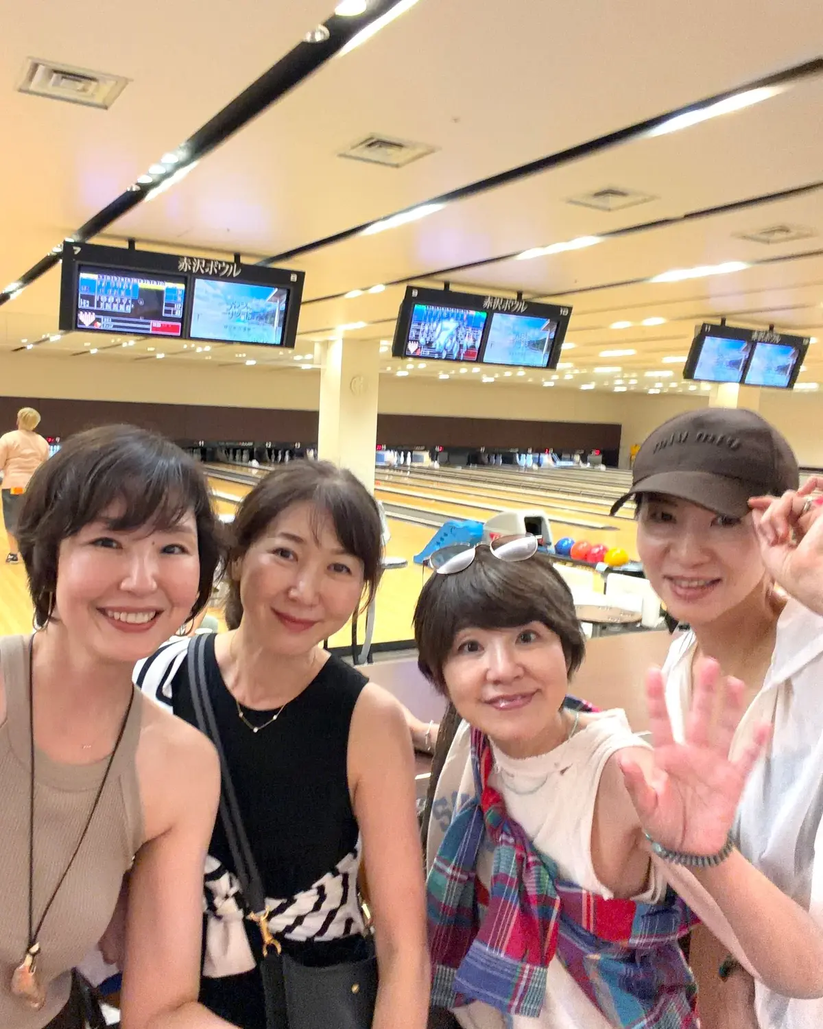 ボウリング場で4人の女性が笑顔で写っている。中央にボウリングボールを持ち、背景にはレーンとスコア表示モニターが見える。
