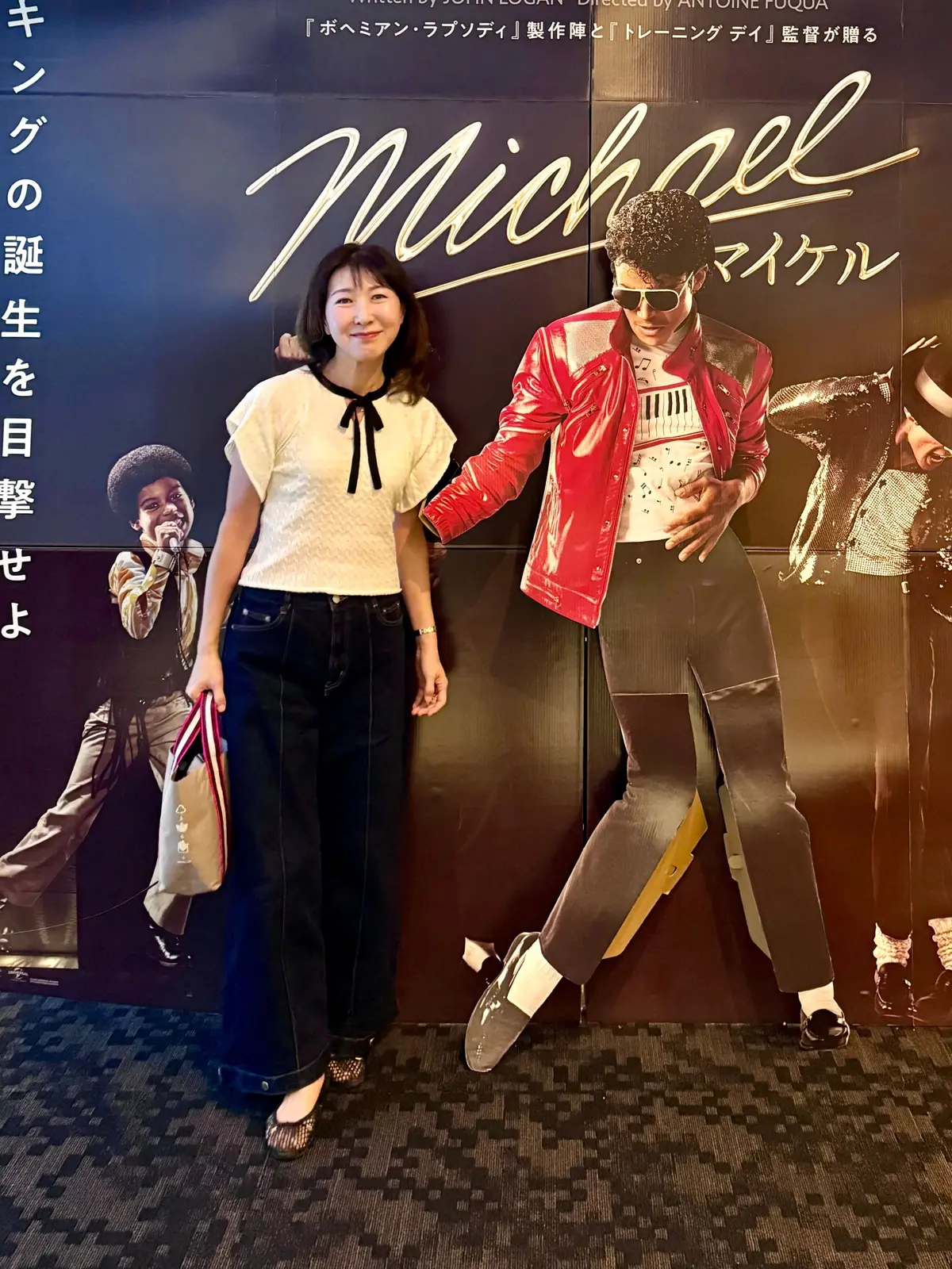 映画「マイケル」のポスターの前で、女性が笑顔で立っている。女性は黒い服に白いスカーフを着用し、隣にはマイケル・ジャクソンの姿が写ったポスターがある。