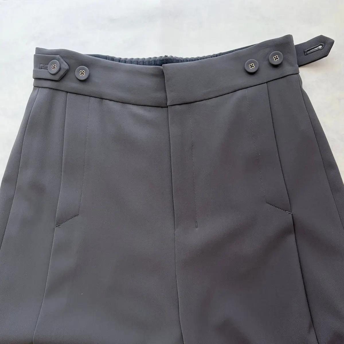 濃いグレーのテーパードパンツ。フロントにプリーツ、右側にフラップポケット。ウエストには3つのボタンが付き、裏地は黒。