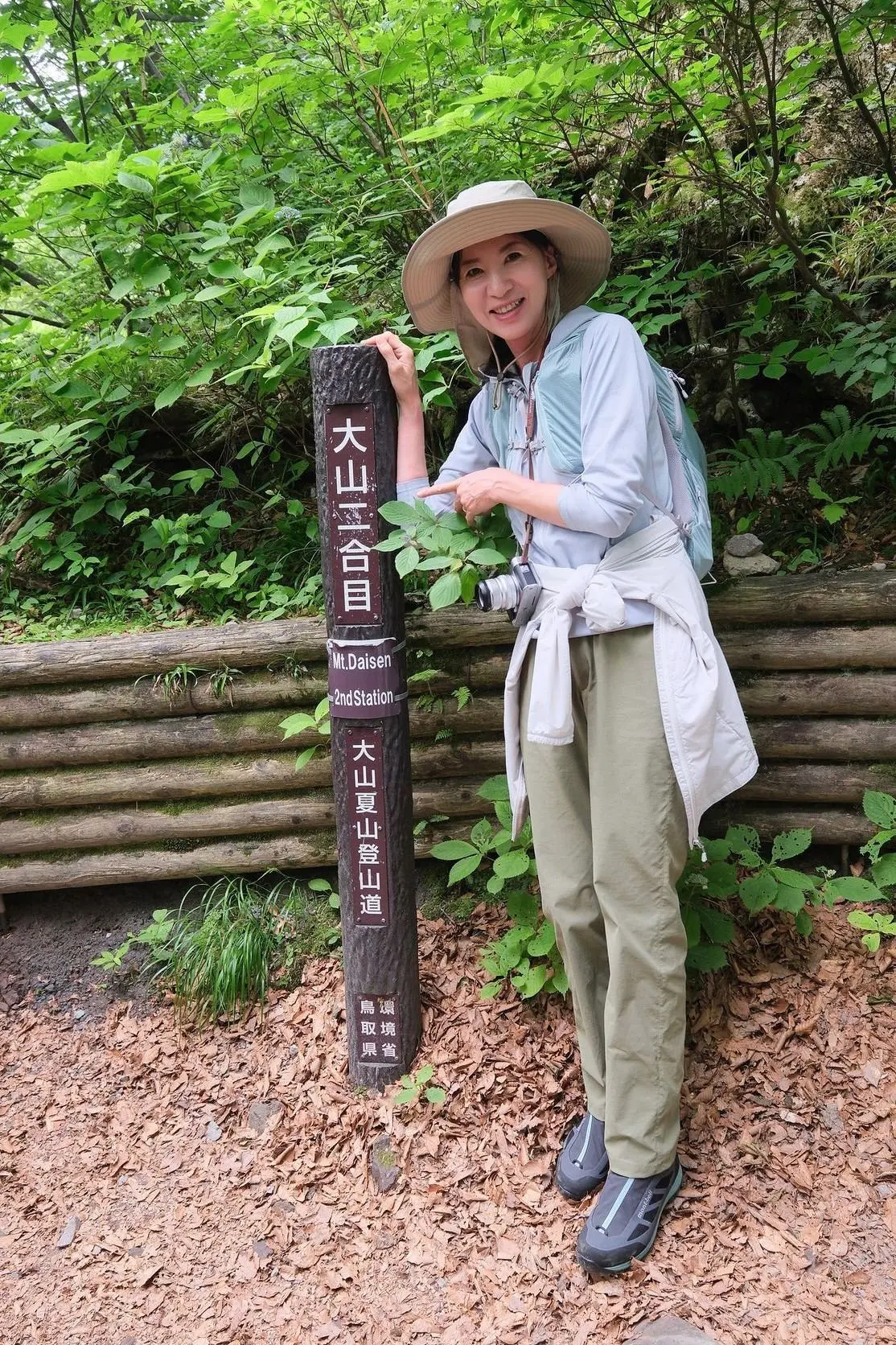 女性が山道で登山標識を指さす。広葉樹の緑豊かな森が背景で、彼女は麦わら帽子とリュックを背負っている。