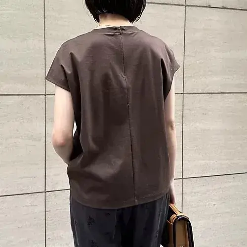 茶色の無地Tシャツを着用した女性。背中心に縦の縫い目があり、袖は短く、肩幅が広め。黒いスカートを履き、茶色のバッグを持つ。背景はグレーのタイル壁。