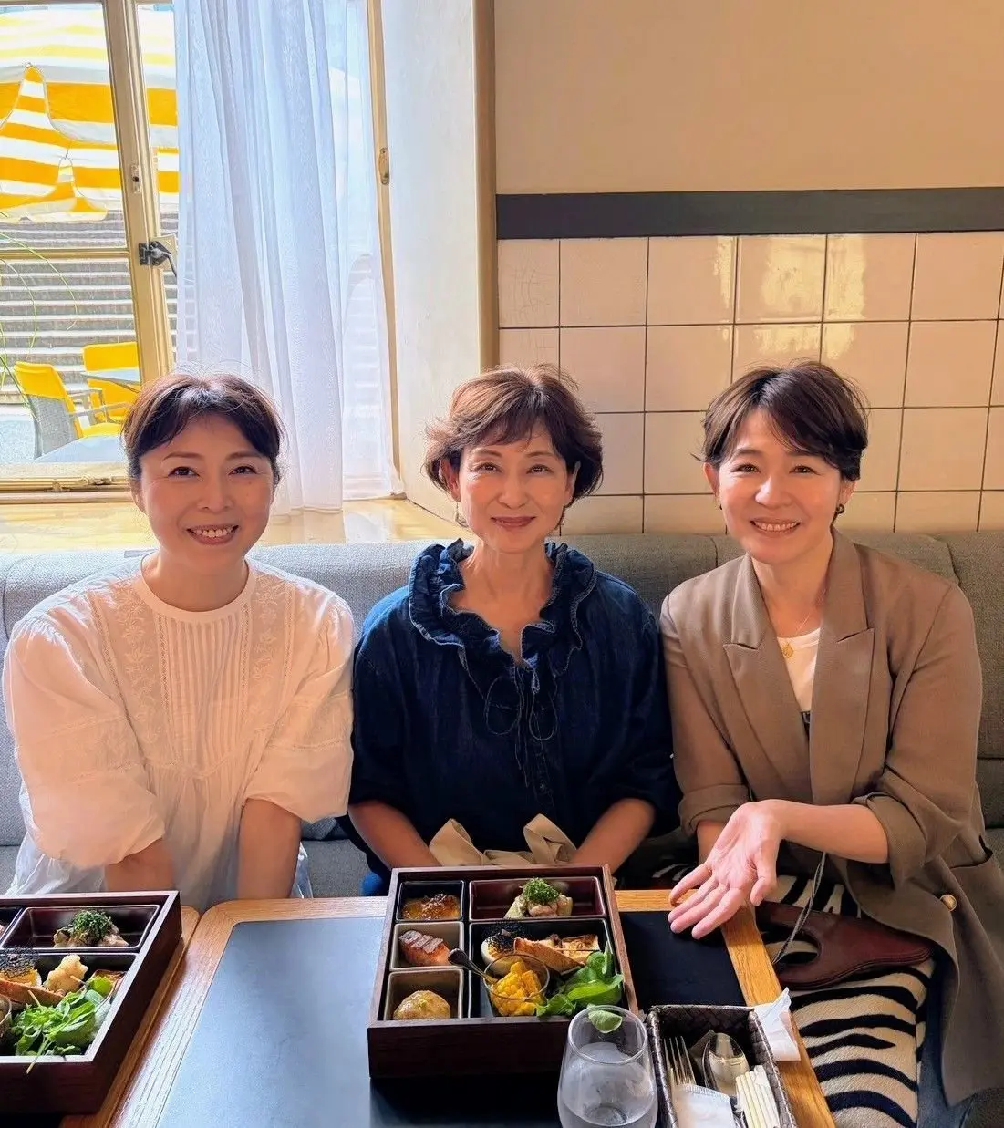笑顔の3人組。中央の女性は青い服、左右の女性は白とグレーの服を着ている。木製のテーブルには、お弁当のような箱に入った料理が並んでいる。背景には窓と白いタイルが見える。