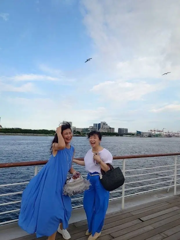 水辺に立つ2人の女性。左は青いロングワンピース、右は白のトップスに青いパンツ。笑顔でこちらを見ている。背景に水面と都市の風景、青空に鳥が飛んでいる。