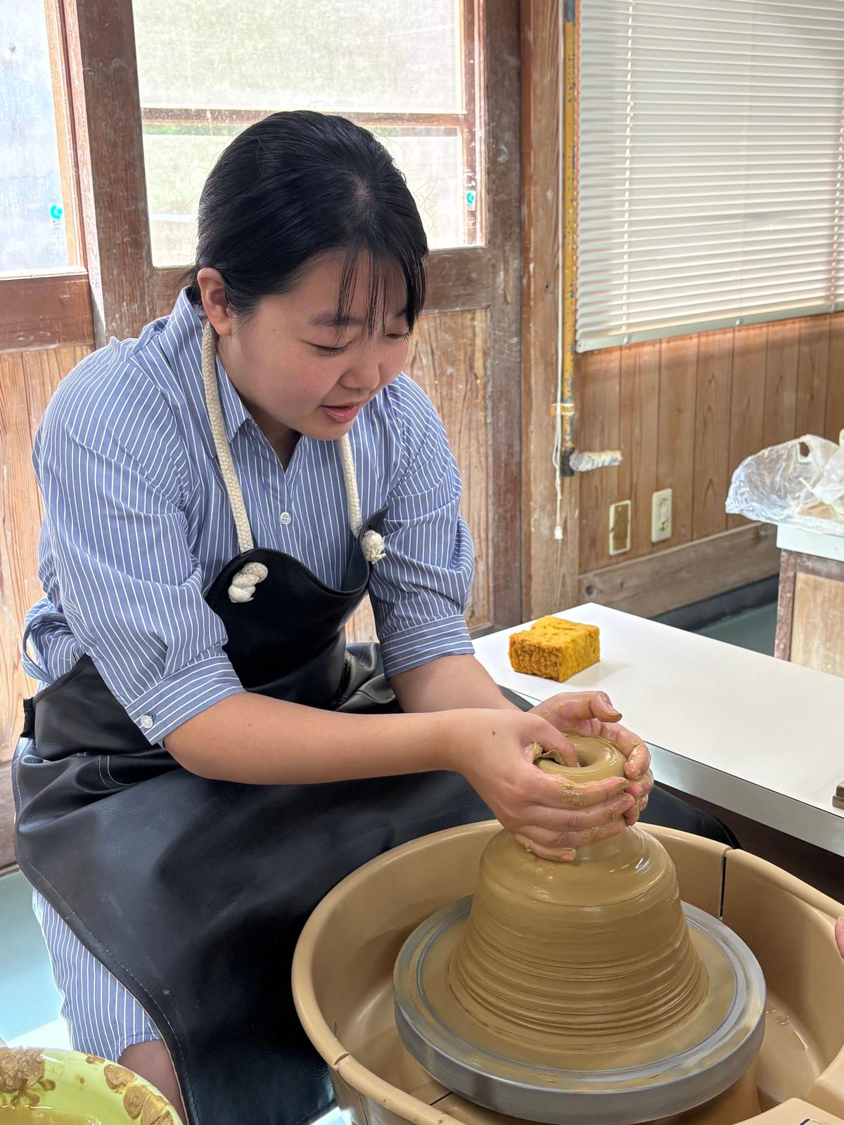 女性が陶器の電動ろくろで土台を成形している。手で粘土を整え、作業台には黄色の焼き物と工具が置かれている。